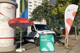 Infostand Digtialer Engel mit Litfa&szlig;s&auml;ule, Sonnenschirm, Tresen. Infomobil im Hintergrund.