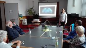 mehrere Personen sitzen mit Abstand um zusammensgestellte Tische und schauen auf eine Pr&auml;sentation auf eienr Leinwand