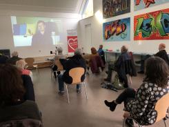 Personen sitzen mit Abstand auf St&uuml;hlen in einem Raum und schauen Video auf Leinwand
