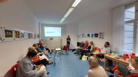 Blick in einem Raum, in dem rund 14 St&uuml;hle mit Senior:innen besetzt sind. Sie lauschen einer Vortragenden. 