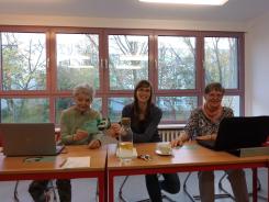 Die Referentin sitzt mit zwei Teilnehmerinnen an einer Tischreihe und l&auml;cheln in die Kamera. Vor beiden Teilnehmerinnen stehen Laptops. 