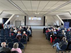 Viele Teilnehmenden sind von hinten zu sehen. Sie sitzen auf St&uuml;hlen und blicken nach vorne, wo auf einer Leinwand eine Videokonferenz zu sehen ist.