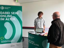 Mobilreferent Johannes ist im Gespr&auml;ch mit einem Fachbesucher am Messestand des Digitalen Engel. Auf der linken Seite steht ein Roll Up mit der Aufschrift "Dabei sein! Online im Alter."