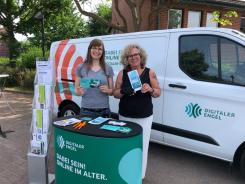 Petra vom Digitalen Engel und Kooperationspartnerin Frau Andermann stehen am Info-Stand vor dem Info-Mobil und halten zwei Flyer. Sie l&auml;cheln in die Kamera.