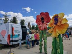 Zwei als &uuml;bergro&szlig;e Blumen verkleidete Maskottchen des Landesseniorentages stehen neben unserem Infomobil an dem sich einige Besucherinnen mit Johanna unterhalten 