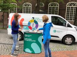 Petra steht mit Melanie Sing von der Stadt Elmshorn am Gl&uuml;cksrad vor dem Info-Mobil. Frau Sing dreht das Gl&uuml;cksrad und Petra h&auml;lt Zettel mit Fragen in der Hand.