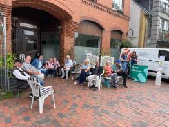 Das Info-Mobil steht vor dem Caf&eacute;. Petra bespricht mit einer Seniorin ein individuelles Thema. Daneben sitzen viele Senior:innen auf St&uuml;hlen und trinken Kaffee.