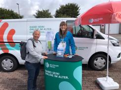 Frau D&uuml;&szlig;mann und Petra stehen am Info-Stand vor dem Info-Mobil und l&auml;cheln in die Kamera.