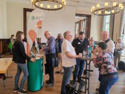 Petra steht am Info-Stand des Digitalen Engel w&auml;hrend der Markt der M&ouml;glichkeiten und ist im Gespr&auml;ch mit einem &auml;lteren Herrn. Neben dem Stand stehen vier &auml;ltere Menschen um einen runden Tisch und unterhalten sich. 