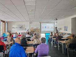Die Teilnehmenden sitzen an Tischen im Raum und blicken zur Pr&auml;sentation an der Leinwand