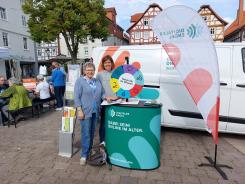 Theresa und eine Besucherin l&auml;cheln in die Kamera, links neben unserem Mobil sitzen einige Senior:innen an einem Tisch auf 2 Partyb&auml;nken.