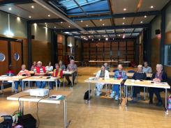 Blick von vorne in den Raum auf die rund 15 Teilnehmenden der Veranstaltungen. Sie sitzen auf St&uuml;hlen an Tischen, auf denen Unterlagen und Smartphones liegen.