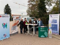 F&uuml;nf Personen stehen vor dem Info-Mobil und halten Flyer in die Kamera. Um sie herum stehen drei verschiedene Roll-Ups.