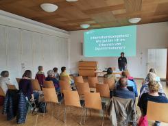 Die Teilnehmenden sitzen auf St&uuml;hlen im Raum und blicken zur Wand nach Vorne