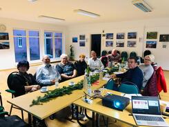 Gruppenbild der Teilnehmenden. Acht Damen und Herren sitzen an einem winterlich geschm&uuml;cktem Tisch. Im Vordergrund sind ein Laptop und ein Beamer zu sehen.