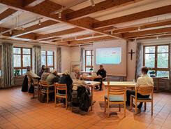 Die Teilnehmenden sitzen an Tischen nim Raum und blicken zur Pr&auml;sentation an der Leinwand