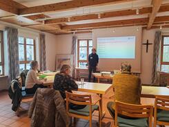 Die Teilnehmenden sitzen im Raum und blicken zur Pr&auml;sentation an der Leinwand und zu unserem Referenten Johannes der neben der Leinwand steht