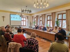 Die Teilnehmenden sitzen an einem gro&szlig;en Tisch im Sitzungssaal und schreiben