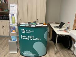 Der Prospektst&auml;nder und der Tisch sind mit Materialien gef&uuml;llt und stehen vor einem Schrank und links neben einem Tisch.