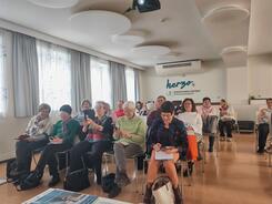Die Teilnehmenden sitzen auf St&uuml;hlen im Raum und schauen zur Pr&auml;sentation an der Leinwand