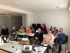 Zw&ouml;f Teilnehmende sitzen auf St&uuml;hlen und blicken nach vorne. Am vorderen Bildrand sieht man einen Laptop und einen Beamer.