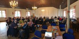 Ein Panorama der knapp 50 Teilnehmenden, die in einem gro&szlig;en Raum an verschiedenen Gruppentischen sitzen. Im Vordergrund steht ein Laptop.