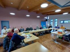 Rund 15 Senior:innen sitzen an Tischen, die in U-Form angeordnet sind. Sie gucken nach vorne auf eine Pr&auml;sentation.