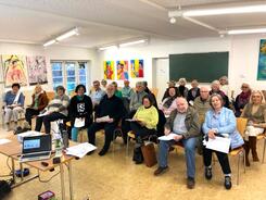 24 der knapp &uuml;ber 30 Teilnehmenden sind auf dem Bild zu sehen. Sie sitzen auf St&uuml;hlen und gucken in die Kamera nach vorne. Einige halten Unterlagen in den H&auml;nden. Im vorderen Bildabschnitt steht ein Laptop und ein Beamer.