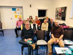 Elf Senior:innen sitzen auf St&uuml;hlen und blicken nach vorne in die Kamera. 