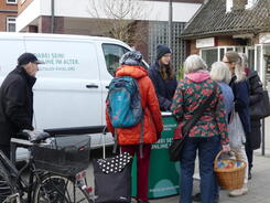 Pera steht in Winterjacke und M&uuml;tze vor dem Info-Mobil am Info-Stand. Um den Stand herum stehen f&uuml;nf Damen mit Einkaufstaschen und ein Herr mit einem Fahrrad.