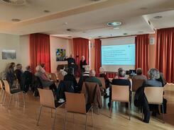 Die Teilnehmenden sitzen im Raum und blicken zur Pr&auml;sentation an der Leinwand und zu Herrn Brockmann, sowie zu einem Mann und einer Frau die neben der Leinwand stehen