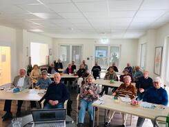 Ca. 20 Senior:innen sitzen in einem Raum vor Tischen. Einige gucken nach vorne in die Kamera. Einige haben Smartphones in den H&auml;nden. Auf den Tischen liegen Info-Materialien.