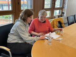 Theresa sitzt mit einer Besucherin am Tisch und erkl&auml;rt Dinge am Smartphone.
