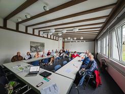 Die Teilnehmenden sitzen an einem gro&szlig;en, zusammengeschobenem Tisch im Raum und blicken in die Kamera