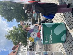 Theresa und eine Besucherin unterhalten sich am Infostand. Hinter ihnen sind ein Brunnen sowie H&auml;user und zwei B&auml;ume zu sehen.