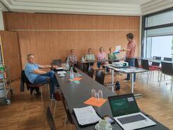 Ein Mann und vier Frauen sitzen an zusammengeschobenen Tischen und blicken zu Johannes, der eine Brosch&uuml;re hochh&auml;lt 