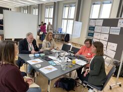 Theresa und die Besuchenden sitzen um einen viereckigen Tisch. Theresa erkl&auml;rt Ihnen Digitale Gesundheitsanwendungen.