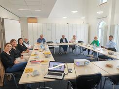 Mehrere Personen sitzen an einem gro&szlig;en zusammengeschobenen Tisch im Raum und blicken nach Vorne 