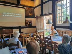 Die Teilnehmenden sitzen an Tischen im Raum und blicken zur Pr&auml;sentation an der Leinwand vorne