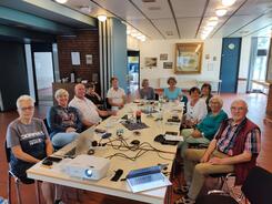 Elf Senior:innen, daovn neun Frauen und zwei M&auml;nner, sitzen an einem gro&szlig;en langen Tisch und gucken nach vorne in die Kamera,