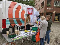 Theresa und eine Besucherin, die links neben ihr steht, sind von hinten zu sehen und h&ouml;ren einer Caritas-Mitarbeiterin zu, die am gemeinsamen Stand vor dem Infomobil steht.
