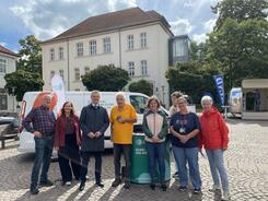 Theresa, die Projektleitung Katharina Kunze, der Abteilungsleiter des BMFSFJ sowie f&uuml;nf Besucher:innen aus Lauenburg stehen auf einem &ouml;ffentlichen Platz vor dem Infomobil und l&auml;cheln in die Kamera.