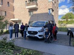 4 Teilnehmerinnen gemischten Alters stehen links neben dem Infomobil sowie drei Teilnehmer und eine Teilnehmerin rechts neben dem Infomobil.  Das Mobil steht auf einer sandigen Fl&auml;che umgeben von einem gepflasterten Weg. Links im Hintergrund ist ein mehrgesch&ouml;ssiges, neueres Haus, rechts im Hintergrund  B&auml;ume, Gras und Wolken. 