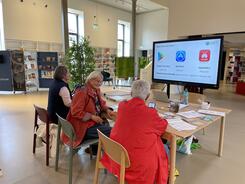 Die drei Teilnehmerinnen sitzen an einem gro&szlig;en Tisch und sind von hinten zu sehen. Die Veranstaltung ist in einem gr&ouml;&szlig;eren B&uuml;chereisaal. Vorne ist die Pr&auml;sentationsfolie zum Thema App herunterladen auf einem Smartboard abgebildet.