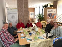Sechs Senior:innen (f&uuml;nf M&auml;nner und eine Frau) und eine Ansprechpartnerin von PiA sitzen an einem Tisch im Kreis und gucken nach vorne in die Kamera.