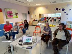 Vier Seniorinnen und ein Senior sitzen auf St&uuml;hlen und blicken in die Kamera. Zwischen den Personen steht ein Tisch mit einem Laptop darauf.