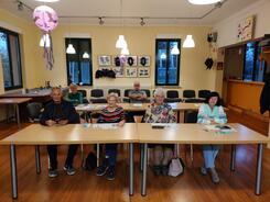 Im Veranstaltungsraum stehen Tische in zwei Reihen. In der ersten Reihe sitzen vier Seniorinnen und ein Senior (links). In der zweiten Reihe sitzt eine Seniorin (links) und ein Senior (miittig). Sie blicken alle nach vorne in die Kamera. Einige l&auml;cheln.