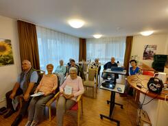In einem Raum sitzen neun Seniorinnen und Senioren auf St&uuml;hlen ohne Tische. Rechts im Bild stehen ein Beamer und ein Laptop.
