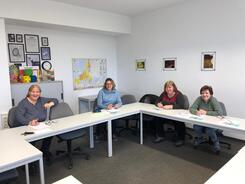 Vier Frauen sitzen an Tischen in U-Form und l&auml;cheln in die Kamera. 