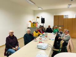 Zehn Senior:innen, darunter zwei M&auml;nner, sitzen an einem langen Tisch und gucken nach vorne in die Kamera. Auf dem Tisch liegen Informationsmaterialien. Vorne auf dem Tisch steht ein Beamer.